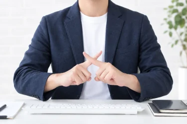 デスクで両手の人差し指を交差させ、拒否のジェスチャーをするジャケット姿の人物