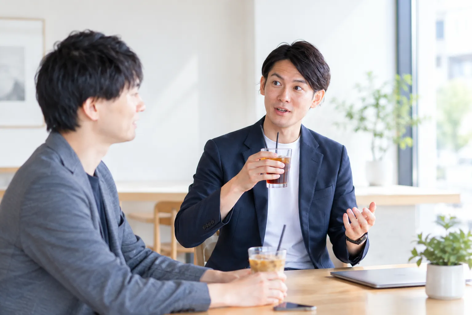 カフェのテーブルで飲み物を前に、打ち解けた様子で仕事の話をする二人のビジネスパーソン